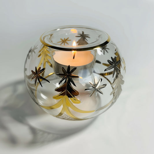 A hand blown glass candle holder with gold and silver Christmas tree decorations and a lit tea light candle inside.
