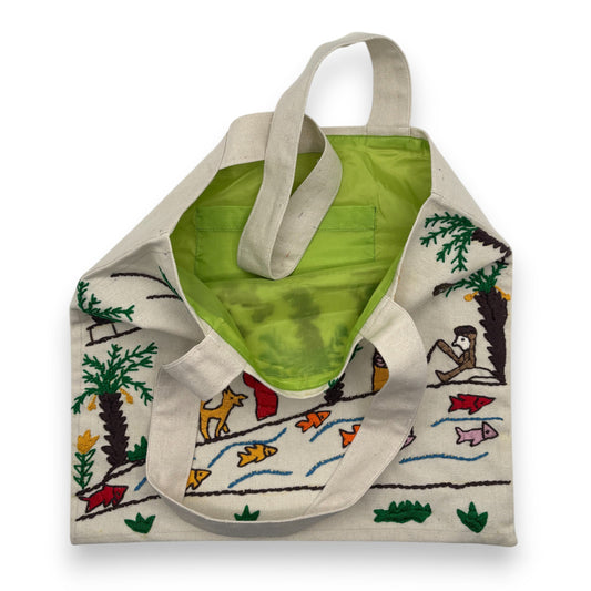 Off-white canvas tote bag with hand-embroidered nature-themed design and green interior lining with pocket